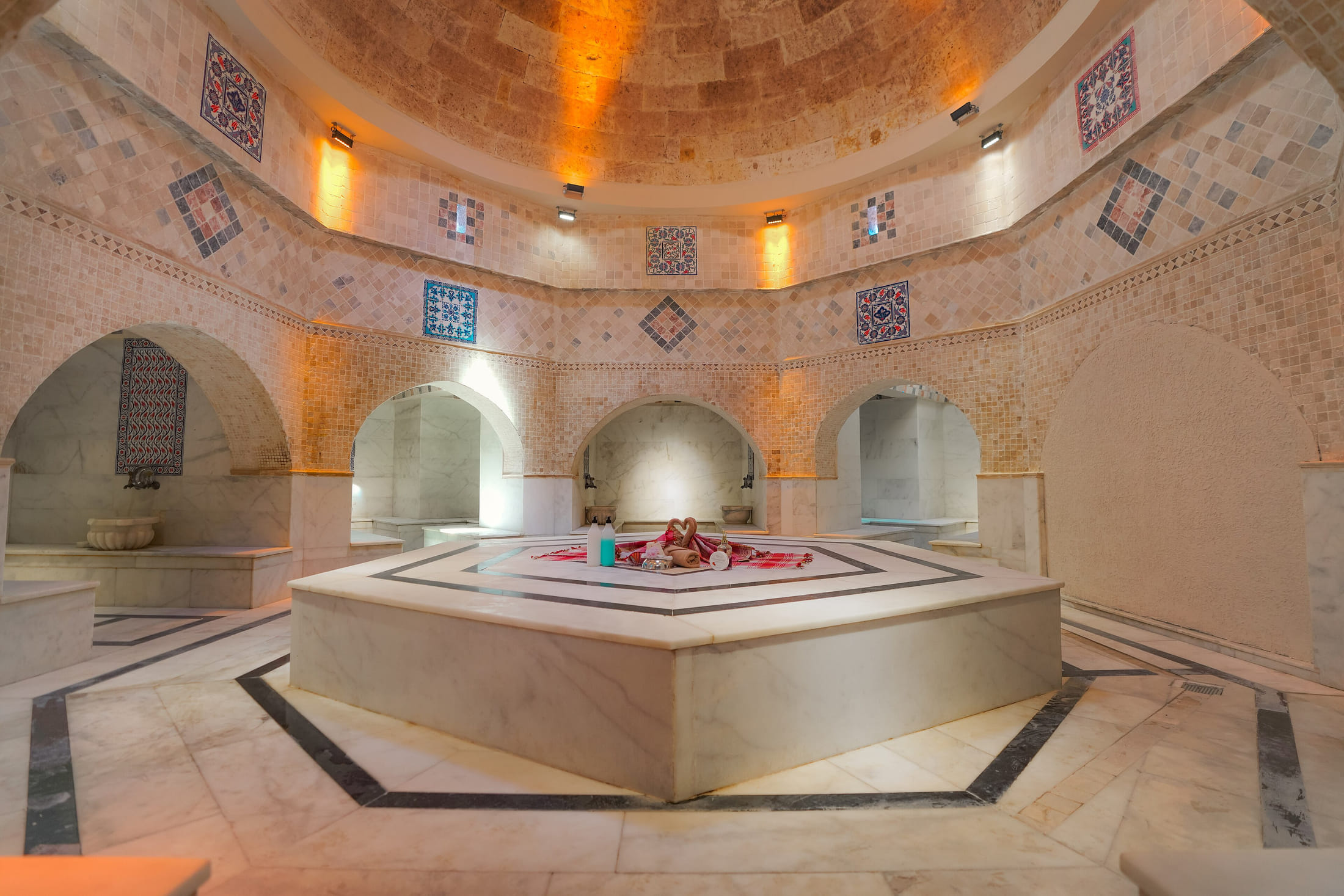 Salle chaude d'un bain traditionnel : pierre claire, niches en arc abritant des vasques de marbre, grande pierre centrale polie et coupole percée au-dessus.