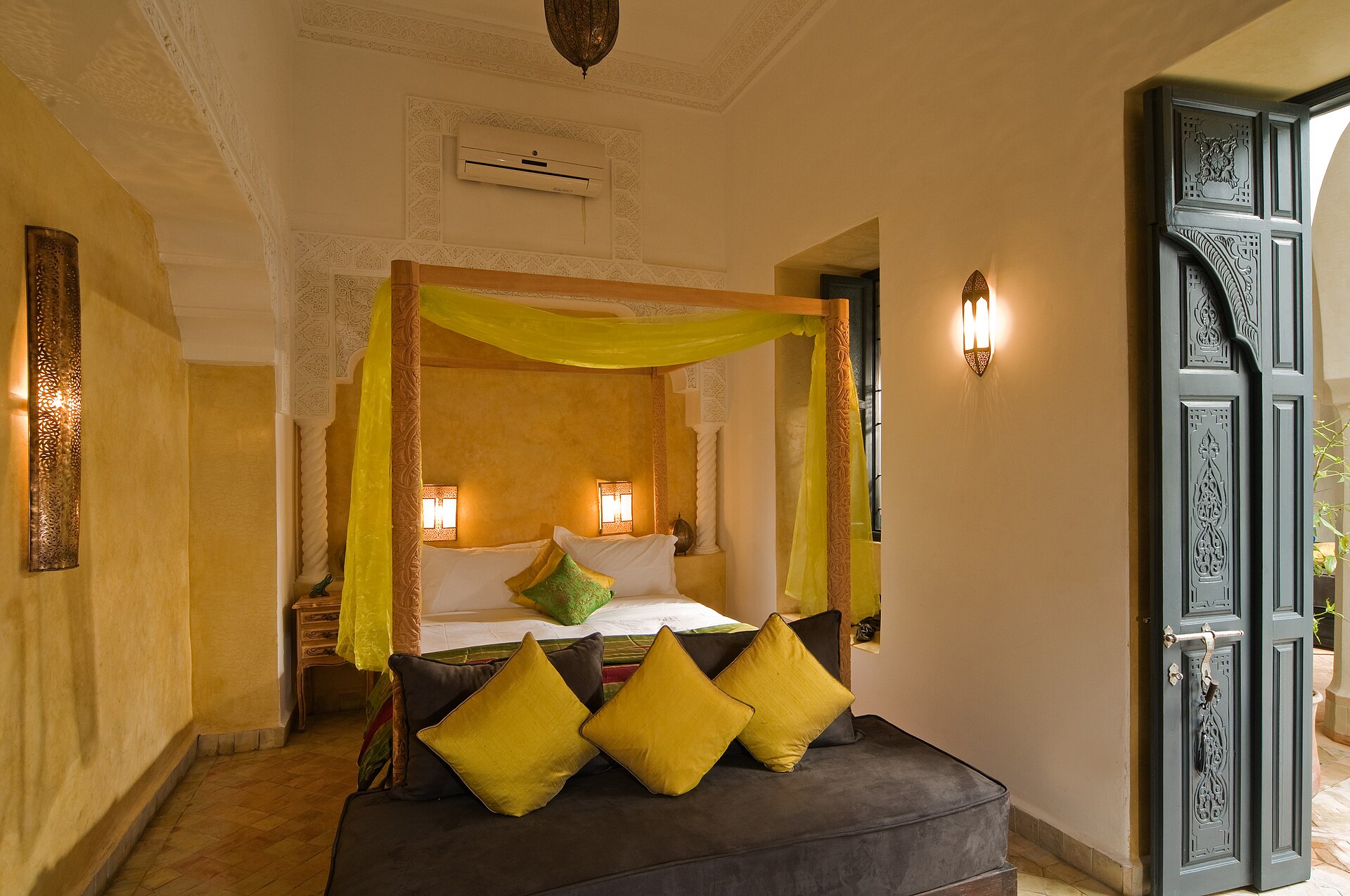 Chambre marocaine aux murs de tadelakt ocre : lit à baldaquin de bois drapé d'un voile jaune safran, appliques de métal ajouré, porte de cèdre sculptée ouverte sur la lumière.