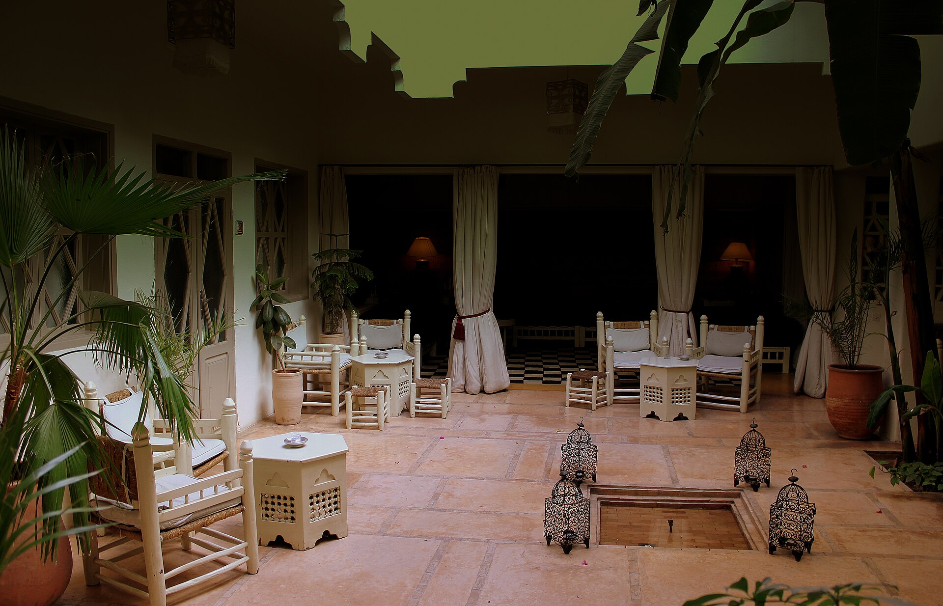Cour de riad au crépuscule : tables dressées de blanc entre les colonnes, fauteuils bas de bois clair, lanternes posées au sol autour d'un bassin et bananiers en bordure.