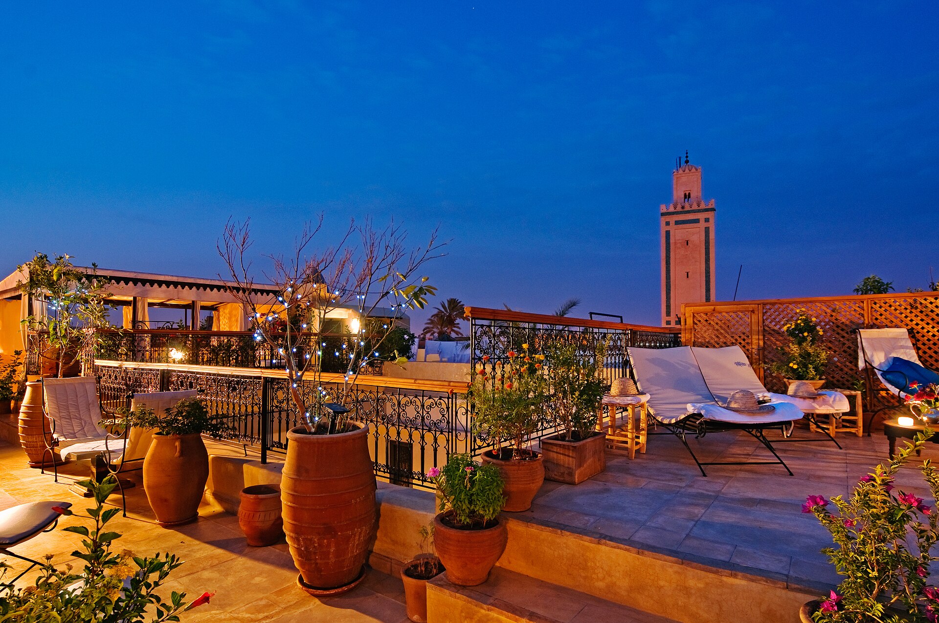 Terrasse sur les toits d'une médina à l'heure bleue : transats de bois, jarres et orangers en pot, garde-corps de fer forgé et minaret éclairé sur un ciel bleu nuit.