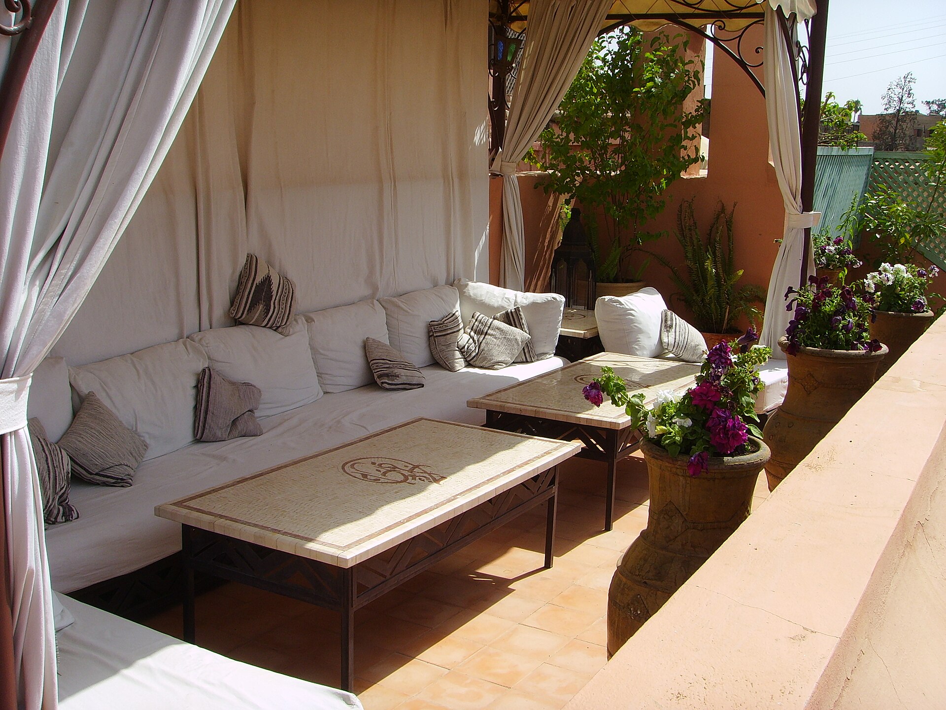 Terrasse de toit ombragée par des voilages de lin : banquette basse garnie de coussins clairs, table basse gravée, jarres de terre cuite et bougainvillier en fleurs.