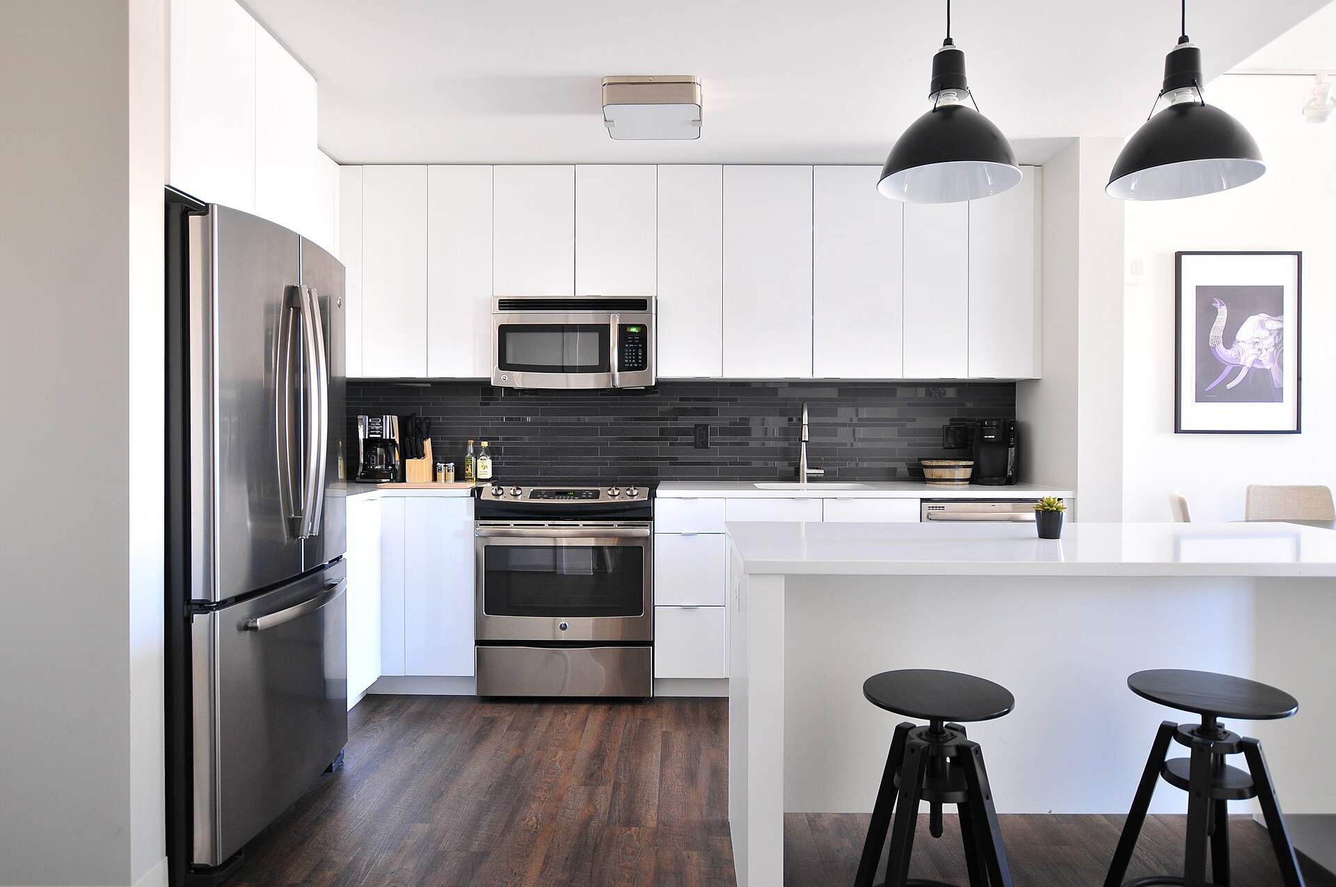 Cuisine contemporaine blanche avec îlot central, crédence sombre et suspensions noires.