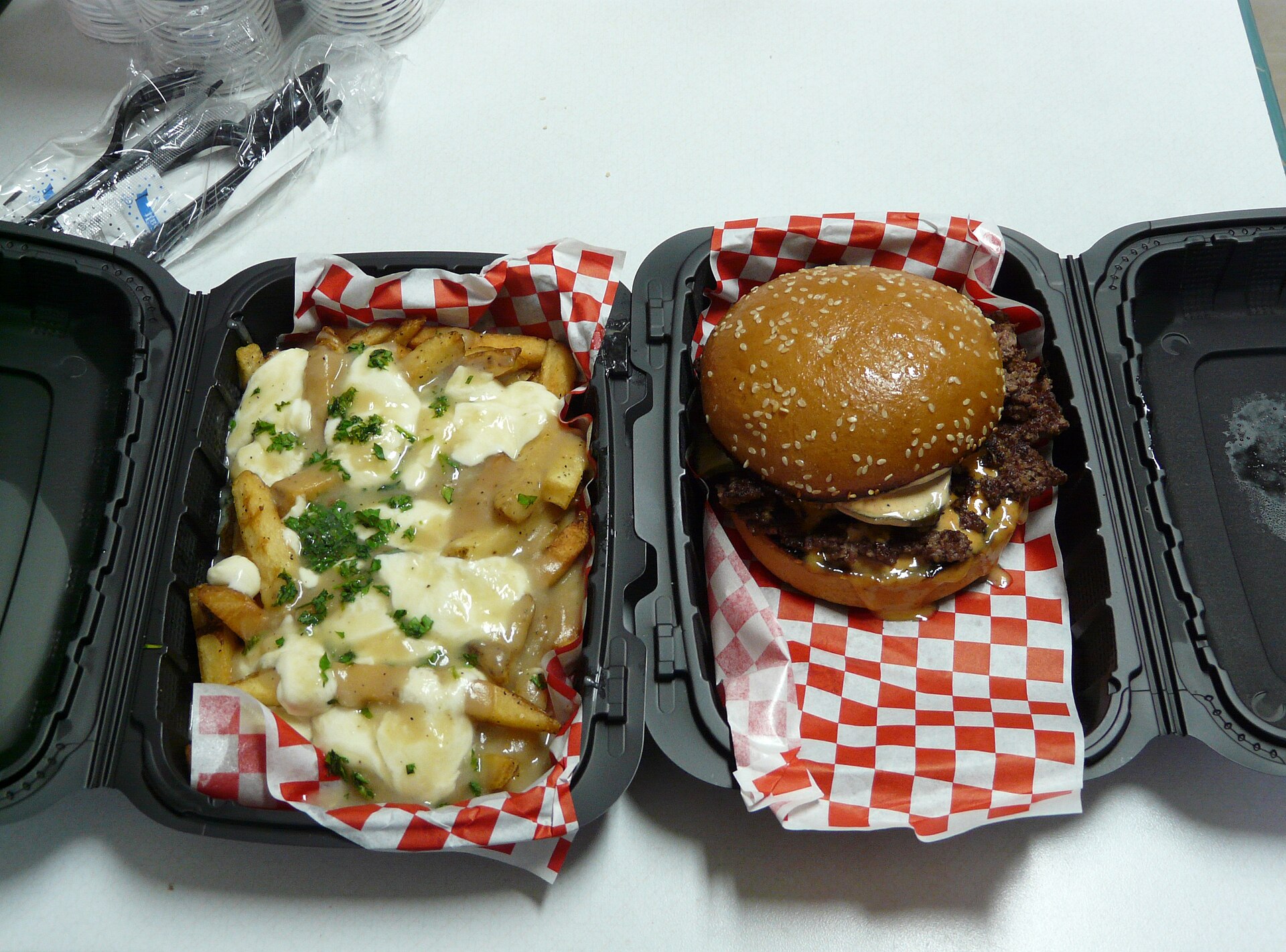 Deux boîtes à emporter ouvertes côte à côte : frites nappées de sauce et de fromage fondu d'un côté, burger smashé sur papier vichy de l'autre.